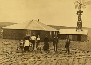 Hembygdsfamilj, utanför sitt torvhus med en väderkvarn på taket av den angränsande byggnaden i Custer County, Nebraska, ca 1885 (s/v foto) av Solomon D. Butcher