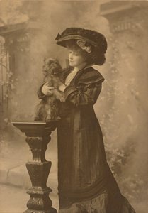 [Sarah Bernhardt står i en studiomiljö och håller i en liten svart hund på en utsmyckad, snidad träpiedestal] av Theodore C. Marceau