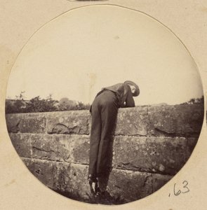 [Man i hatt böjd över en stenmur] av Unknown artist