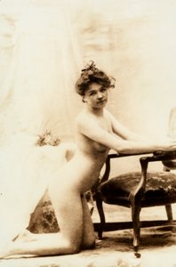 Nakenporträtt av en ung kvinna som står på knä framför en liten fåtölj. av Unknown photographer