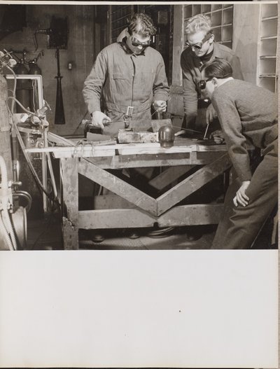 [Tre män med skyddsglasögon svetsar en metallbit på ett bord] av Arnold Eagle