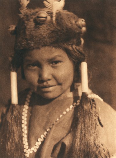 Närbild av en indianpojke från Klamath-stammen eller -byn. Han bär en pälsmössa med påsatta ekollon och fjädrar och har håret uppsatt i två hästsvansar av Edward Sheriff Curtis