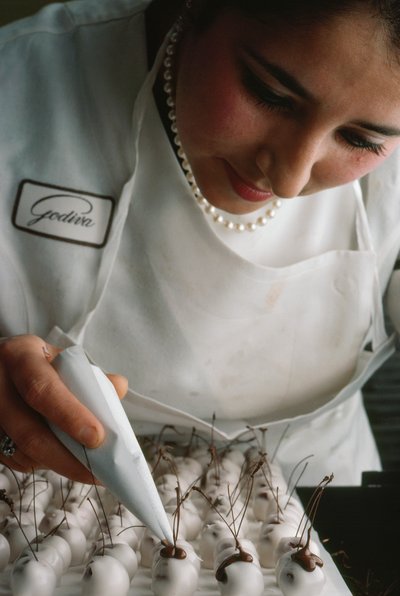 En Godiva-kock fäster chokladstjälkar på fondantöverdragna körsbär. Bryssel, Belgien. av James L Stanfield