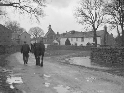 Man leder häst längs väg bort från kamera mot bybyggnader och kyrka; arkitektur; religiös; kyrklig; av Joseph Hardman