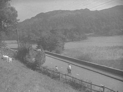Vy över en väg som går längs med Rydal Water, med människor på cyklar, boskap och fjäll av Joseph Hardman