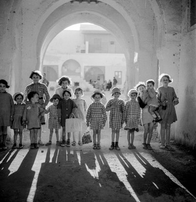 Europeiska barn i skoluniform under båge med skuggor. av Maynard Owen Williams