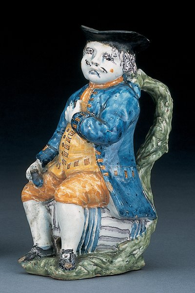 Tobykanna med ansikte och händer i blek mangan, hållande och tom klotformad buff-färgad kanna i tvättad blå jacka och byxor och grön väst, ca 1800 av Unbekannt