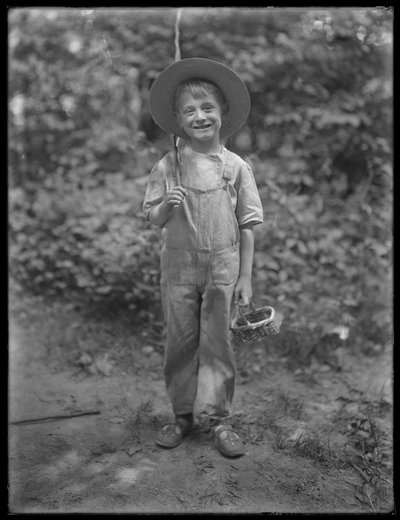 William Grey Hassler i stråhatt och overall, bärande fiskespö och korg i skogen, 20 augusti 1911 av William Davis Hassler