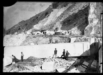 Tre män observerar arbetet med flera grävmaskiner i Apuanska alperna. Bakom dem finns ett enormt block av marmor som redan är avskuret och redo att transporteras till dalen (s/v foto)