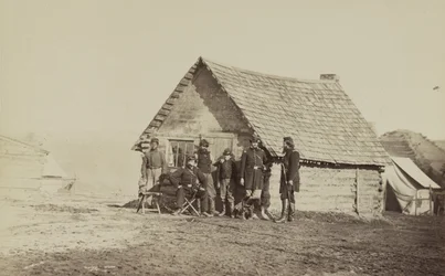 En grupp soldater och två unga män, en afroamerikan, står utanför timmerstugekvarteren, 1861-65