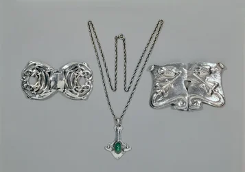 Silversmycken designade för Liberty. Vänster till höger: Midjelås med sammanflätad keltisk design, 1904. Hänge och kedja med cabochonskuren krysopras. Midjelås med repouse-design på hamrad