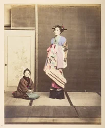 [?Geisha, ] Stående studioporträtt i full längd av en ung kvinna i förhöjda skor, hennes unga skötare på knä på golvet bredvid henne,   Fotograf: Okänd  Lucas Collection: Fotografiskt rekord från Charles James och Morton Ps världsturné, Luc