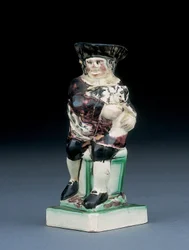 Dick Whittington-kanna, formgjuten reser sig från en stol med hög rygg, håller sin katt, plaskad i bruna, gröna och svarta kläder, en formgjuten sköld vid hans sida, ca 1790