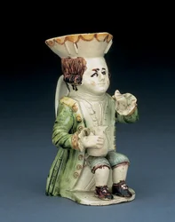 Toby-kanna med en rumlare, hans hatt med brun och gul uddkant, hans inristade bruna hår bundet i stångpiska och med en vit rosett, sittande med en balusterformad kanna på knäet, ca 1770