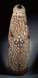 Oval Asmat-sköld, snidad i relief med rullande ornament som innehåller mycket stiliserade figurer, flätade tofsar till bården (trä, raffia och fjädrar)
