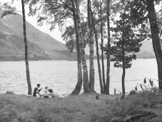 En familj kopplar av nära en sjö med träd till höger och fjäll i bakgrunden