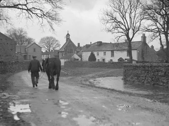 Man leder häst längs väg bort från kamera mot bybyggnader och kyrka; arkitektur; religiös; kyrklig;