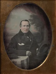 Man som bär kappa och handskar, håller hatt, sitter framför målad utomhusbakgrund, 1840-talet.