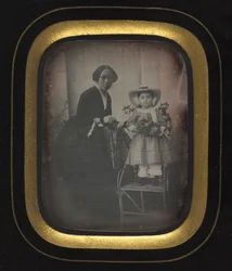 Barn som står på en stol och håller blommor, med mamma, ca. 1855.
