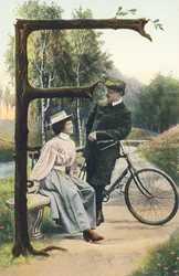 F: En man utklädd till cyklist och lutar sig på sin cykel diskuterar med en kvinna som sitter på en bänk. Landsdekor - Alfabettecken Ref Th. E., L. Theochrom - Serie 994. Theodor Eisman, förläggare, Leipzig