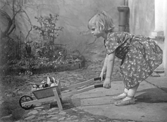 Liten flicka som leker med en skottkärra. Fotografering omkring 1931