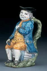 Tobykanna med ansikte och händer i blek mangan, hållande och tom klotformad buff-färgad kanna i tvättad blå jacka och byxor och grön väst, ca 1800