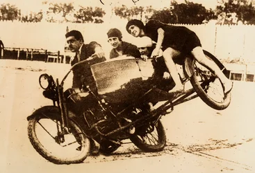En man visar upp sig när han kör en motorcykel med sidovagn som transporterar tre unga kvinnor. Sidovagnen lyfter från marken när de svänger.