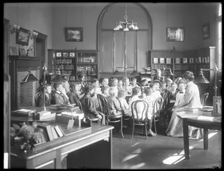Barn lyssnar på en kvinna i läsesalen i en oidentifierad filial av Queens Borough Public Library, omkring 1910.
