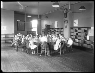 Barn lyssnar på en kvinna i läsesalen på en oidentifierad filial till Queens Borough Public Library, ca 1910