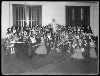 Gruppporträtt av en flickkörsgrupp som samlas runt ett piano med noter i handen, troligen vid det romersk-katolska barnhemmet i Kingsbridge, Bronx, omkring 1911.