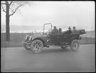Tre oidentifierade personer och två Chow-hundar som kör i en cabriolet bil längs en flod eller sjö, ca 1912