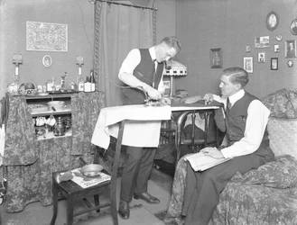 Oidentifierad man som visar användningen av ett elektriskt strykjärn, bevakad av en annan man, ca 1913-14