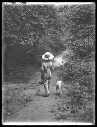 William Grey Hassler i stråhatt och overall, bärande fiskespö och korg, går längs en stig i skogen med en liten hund (Bessie), 20 augusti 1911