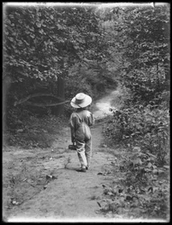 William Grey Hassler i stråhatt och overall, bärande fiskespö och korg, går längs en stig ensam i skogen, 20 augusti 1911