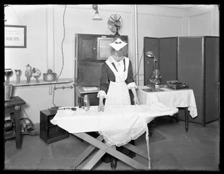 Ung kvinna i en pigadräkt som visar användningen av ett elektriskt strykjärn, ca 1913-14